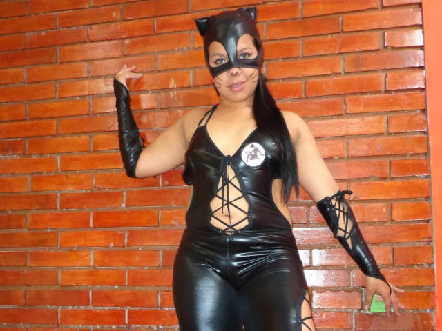 TiaraKitty in black latex cat costume with mask, lace-up front, and arm gloves, showing her tits and ass, standing against a brick wall.