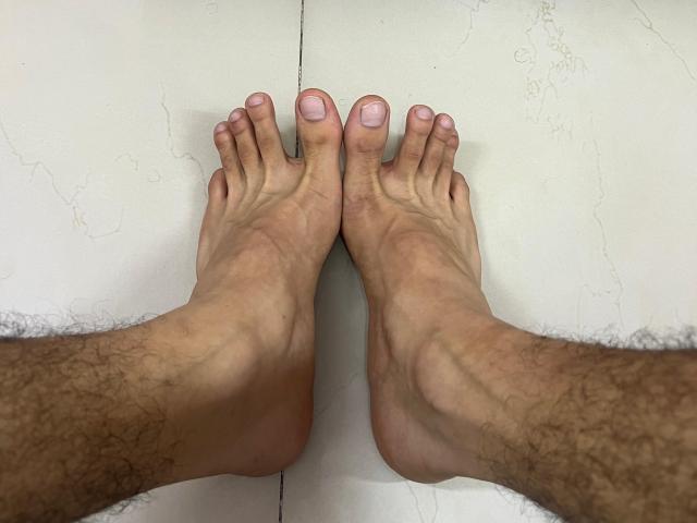 SamuelHills' hairy legs with bare feet, toes pointing upward, pink nail polish on all toes, standing on a white, cracked surface.
