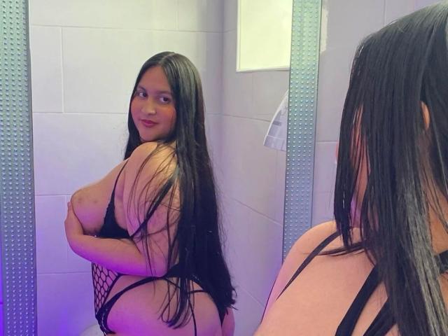MiaCooper1 with long black hair, wearing a black fishnet lingerie, covering her big tits with her right arm, looking at her reflection in a mirror.