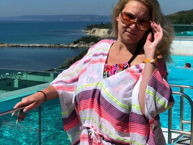 lisabigtits-hot with blonde hair, tan skin, wearing a pink and white striped shawl, orange sunglasses, and a colorful top, standing by a pool, talking on her phone.