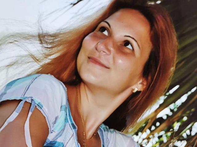 Lisabigtits-hot with light brown hair, smiling slightly, looking up, wearing a blue and white plaid shirt, small gold necklace, and earrings, in a bright, sunny outdoor setting.
