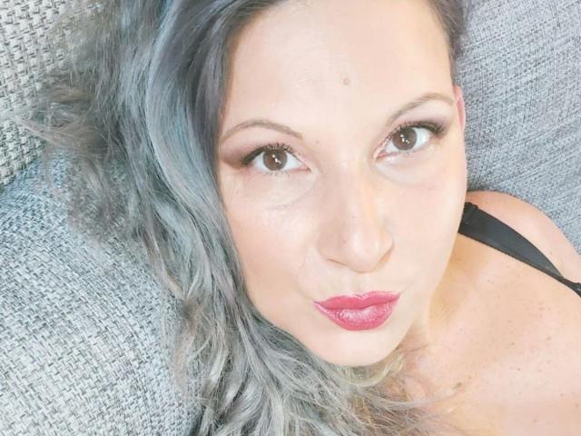 lisabigtits-hot with gray hair, pouty pink lips, and black strap, looking up with brown eyes, lying on a textured gray surface.