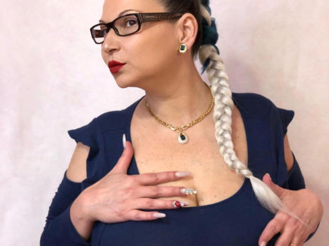 lisabigtits-hot with glasses, red lipstick, blue braided ponytail, gold necklace, holding her tits in a navy top, white background, wearing green earrings and a ring.