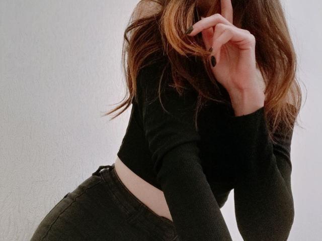 WollyMolly mit langen braunen Haaren, einem schwarzen Crop-Top und dunkler Hose, Hand in der Nähe des Gesichts, zeigt im Bild ihren Hintern..