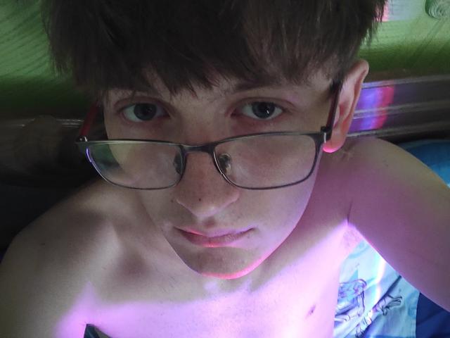 O ShineeGuy usa óculos de nerd, tem sardas e cabelo castanho curto, e está olhando para cima com uma expressão ligeiramente curiosa, sem camisa, com luz rosa e roxa refletindo em seu peito..