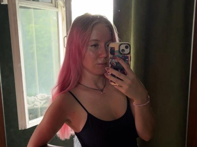Loreen Tracy, con cabello rosa, camiseta negra, sosteniendo un teléfono, selfie frente al espejo, mostrando sus senos, con una ligera mueca, cortina verde, ventana y pulsera en su muñeca..