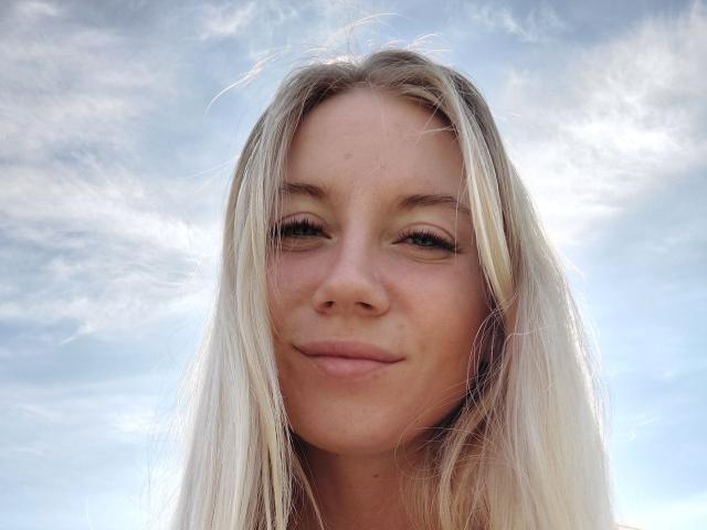 Loreen Tracy, com cabelo loiro, um leve sorriso e maquiagem discreta, tem um rosto bonito e está olhando para a câmera. Seus seios não estão visíveis, apenas sua cabeça e parte superior do peito..