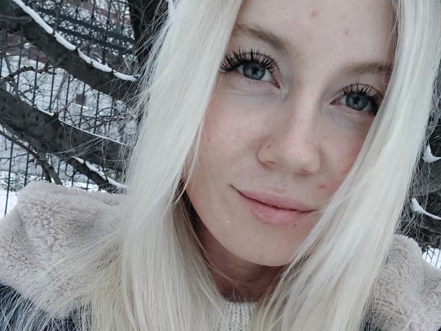 Il viso di Loreen Tracy, con i suoi lunghi capelli bianchi, gli occhi azzurri e le labbra rosa, indossa un cappotto beige soffice. Nella parte inferiore dell'immagine, si vedono rami di alberi coperti di neve. Sono presenti piccole macchie rosse sulla sua guancia..