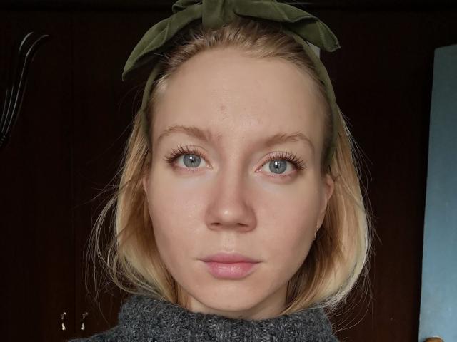 LoreenTracy with blonde hair, green eyes, and a green headband. She's wearing a grey turtleneck, and her face shows a slight zit on her forehead.
