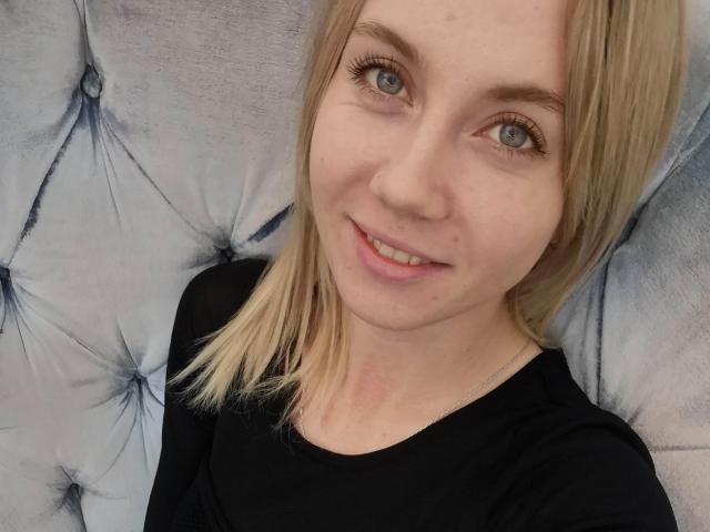 Loreen Tracy, com cabelo loiro, olhos azuis, camisa preta e um sorriso adorável, sentada em um sofá cinza com detalhes em botão. Ela tem um nariz pequeno e não tem tatuagens visíveis..