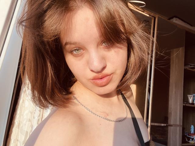 Ssimona has light brown, shoulder-length hair, green eyes, and a slight smile. She's wearing a black strappy top, with a silver necklace, and sunlight highlights her face.