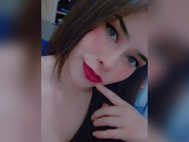 FelicityFate a la peau claire, les yeux bleus, et un blush rose avec du rouge à lèvres rouge. Elle touche son menton avec son doigt, et elle porte du noir. Ses cheveux bruns sont légèrement en désordre..
