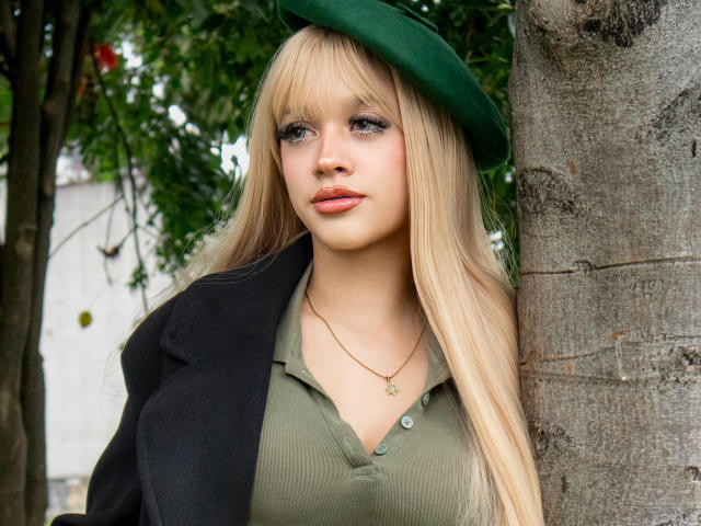 Ada Ferreira mit langen blonden Haaren, einem grünen Hut, einem schwarzen Mantel, einem olivgrünen Hemd, einer Halskette, stehend neben einem Baum, mit grünen Augen, dezenter Schminke, voller Lippen, einem sanften Ausdruck, Baumstamm auf der rechten Seite..