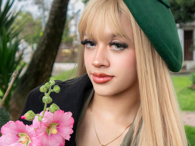 Ada Ferreira, mit blonden Haaren, einer grünen Mütze und rosa Hibiskusblumen in der Hand, hat volle Lippen und Wimpern und trägt eine schwarze Jacke. Sie schaut nach unten und lächelt leicht..