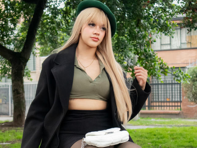 AdaFerreira with long blonde hair, green beret, black coat, olive crop top, black skirt, holding white purse, sitting on grass, green eyes, light makeup, medium tits, outdoors.