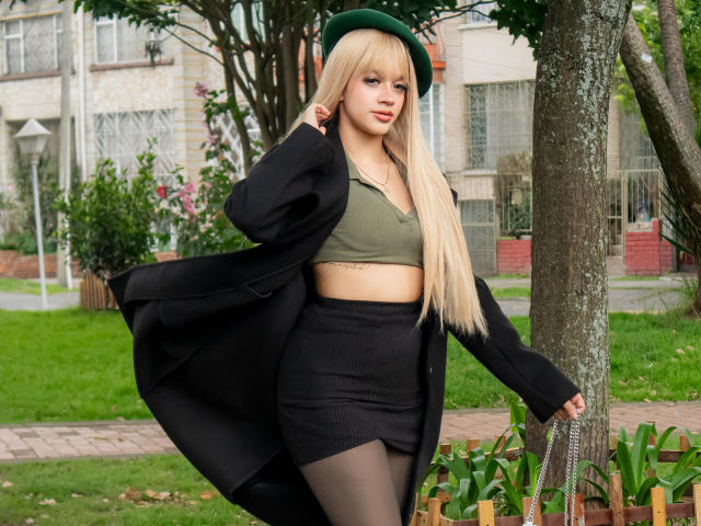 AdaFerreira with long blonde hair, green beret, olive crop top, black skirt, black coat, and sheer pantyhose, standing by a tree, holding a chain purse, looking at camera.
