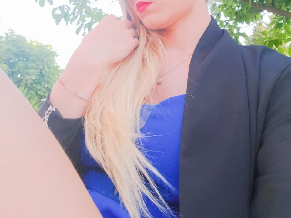 HeleG con capelli biondi, vestito blu, giacca nera, rossetto rosso, mano che tocca i capelli, braccialetto e collana, seduta all'aperto, gambe inquadrate, luce diurna intensa..