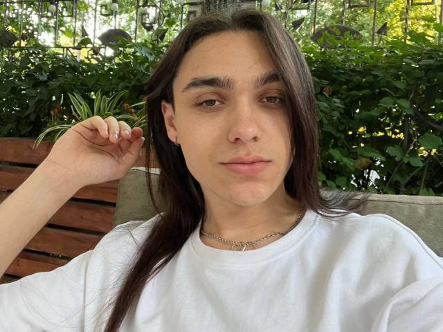 GabiYumy with long brown hair, white tee, and silver necklace, hand in hair, neutral expression, green foliage behind, wooden bench, slightly parted lips, natural light, casual vibe.