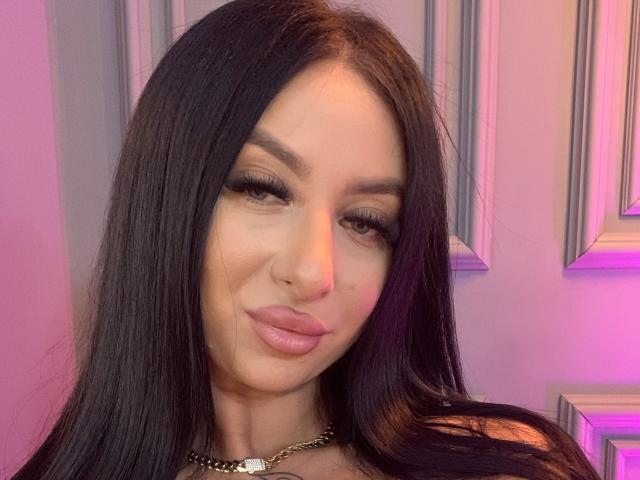 LexyVoss hat langes, schwarzes Haar, volle, rosafarbene Lippen und dichte Wimpern. Sie trägt eine schwarze Halskette mit einem weißen Anhänger und lächelt leicht. Ihre Nase ist gestochen..