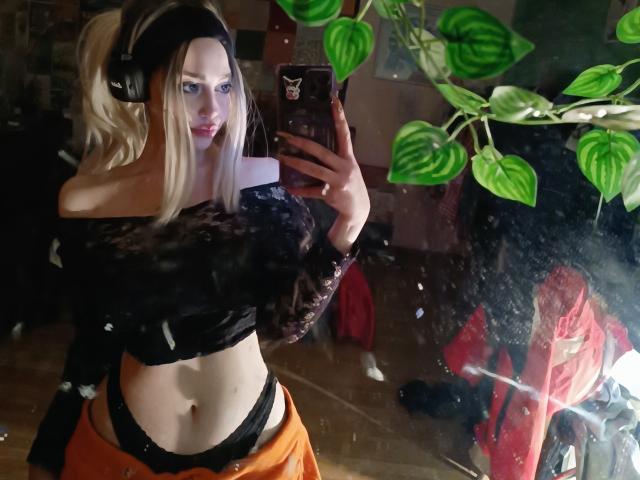 WhiteKitsu with blonde hair, wearing headphones, black off-shoulder top, black underwear, and orange pants, taking a mirror selfie. Green leafy plant in the background.