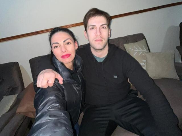DianayJack with red lipstick, black puffy jacket, and black pants, and a brown-haired guy in a black hoodie, sitting on a brown couch, both looking at the camera.