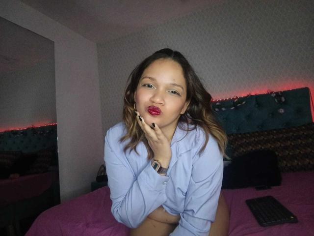 BiancaMorettii with wavy brown hair, pouty red lips, and black nail polish, wearing an unbuttoned light blue shirt, exposing her tits, and a watch, on a pink bed.