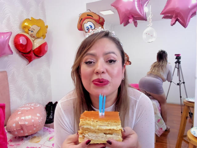 AntonellaRiquel with pink lipstick, holding a slice of cake with blue candles, wearing a white sheer top, brown hair, pink balloons, and a star-shaped balloon in the background.