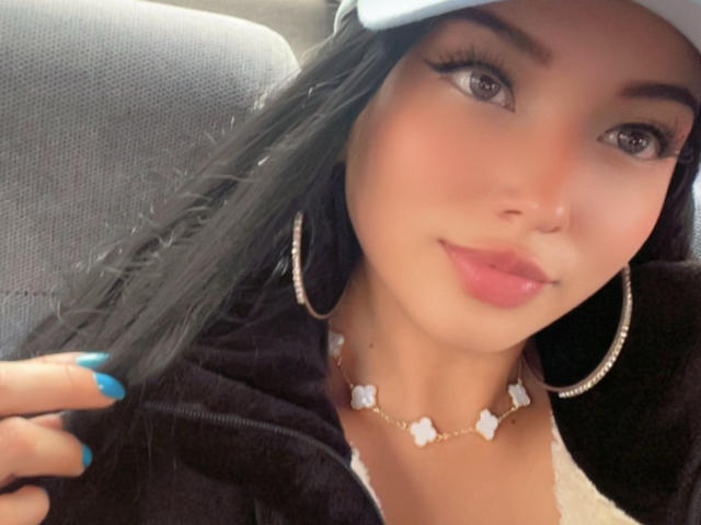 LillyTaylor has long black hair, blue nails, a white cap, hoop earrings, a flower necklace, and pink lips, looking at the camera with a slight smile.