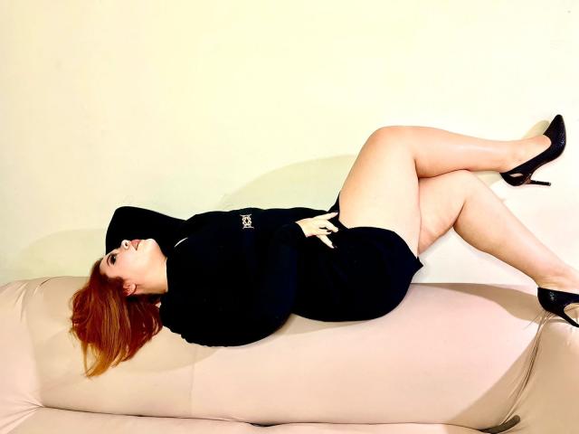 MaddyBlooms is lying on a beige couch, wearing a black dress and black high heels, with her legs crossed, showing her ass and red hair.
