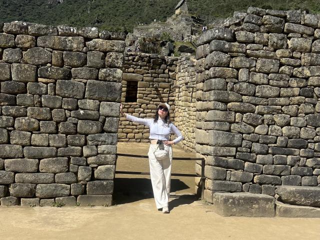 ElizaCanterville stands in a stone ruin, wearing a white shirt, beige pants, and a belt, with her hand on a wall, looking forward, in a sunny, historical setting.