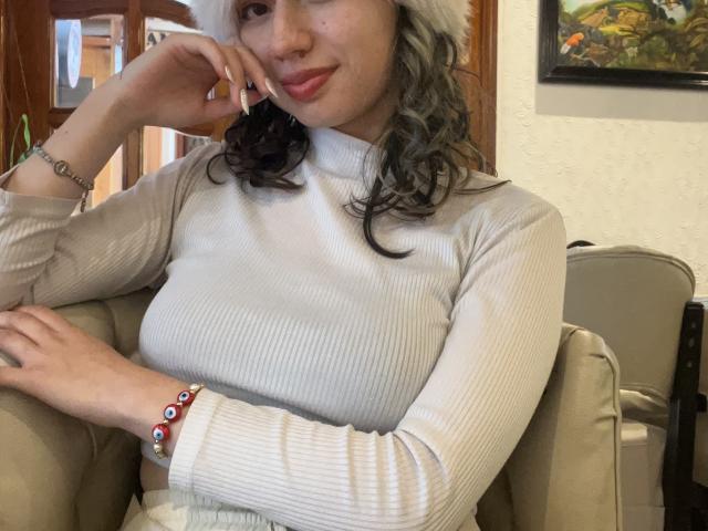 ElizaCanterville with curly gray-black hair, wearing a white ribbed turtleneck, and a white fur hat, resting her chin on her hand, showing off her tits and cute smile.