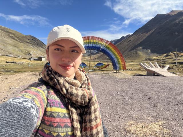 ElizaCanterville with a smile, wearing a white cap, colorful striped sweater, and patterned scarf, standing in front of a rainbow sculpture, mountains and blue sky in the background.