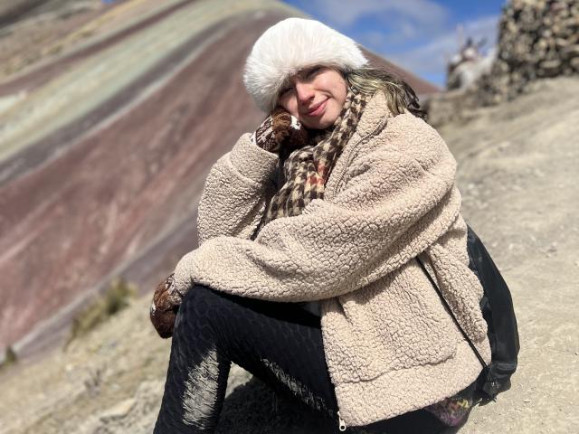 ElizaCanterville is squatting, wearing a fluffy white hat, beige fuzzy coat, brown gloves, and black pants, with a patterned scarf, smiling at the camera, in a rocky landscape.