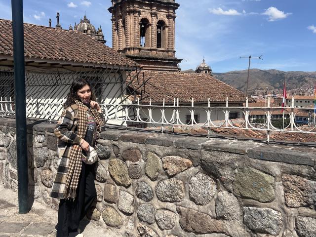 ElizaCanterville stands with a checkered scarf, brown hair, stone wall, and church tower in the background. She's wearing black pants and a brown scarf, looking at the camera.