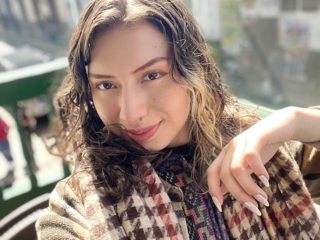 ElizaCanterville with wavy brown hair, a nose ring, and long nails, smiling at the camera, wearing a brown and white checkered coat, and a colorful patterned shirt.