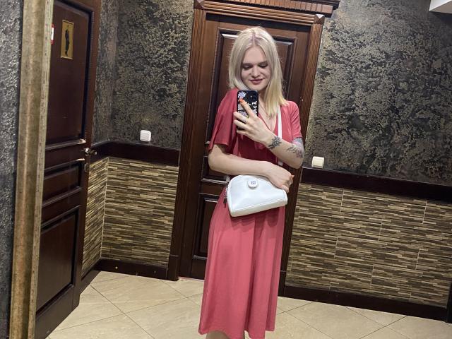 BrendaLigi with blonde hair, pink dress, holding white purse, tattoo on arm, smiling, standing in room, taking mirror selfie with phone, showing her ass and tits.