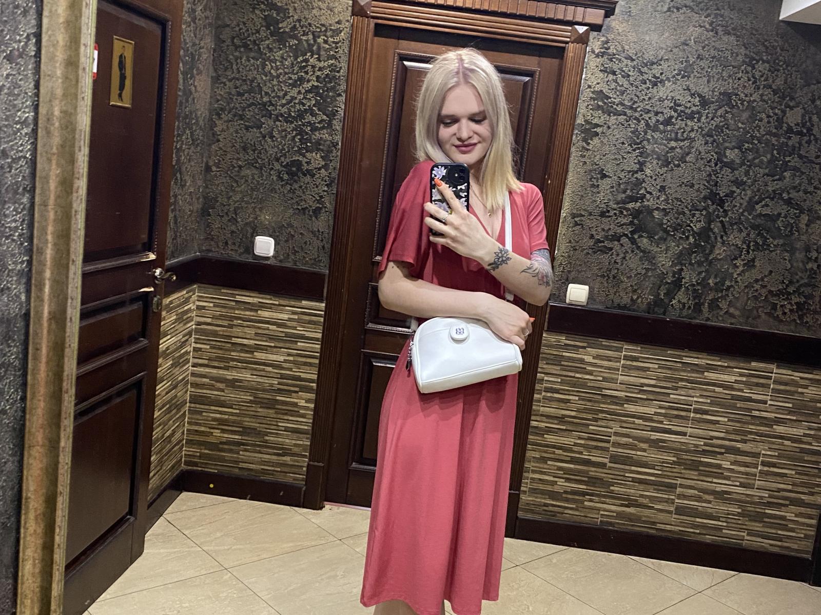 BrendaLigi with blonde hair, pink dress, holding white purse, tattoo on arm, smiling, standing in room, taking mirror selfie with phone, showing her ass and tits.