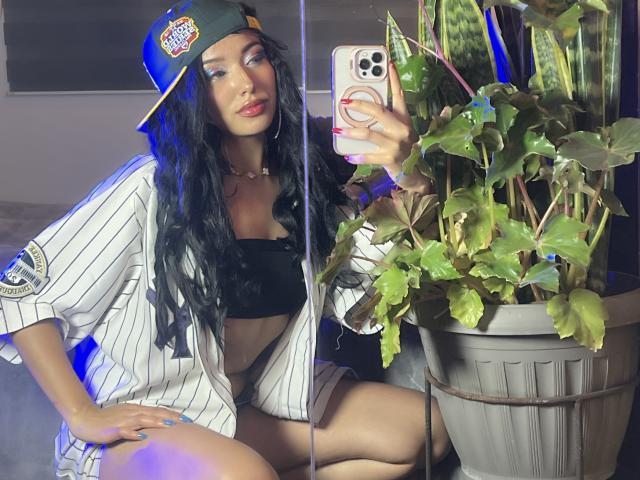 LillyTaylor with long black hair, wearing a baseball cap, pinstripe shirt, black bra, and shorts, taking a mirror selfie, showing her tits, with a plant on the right.