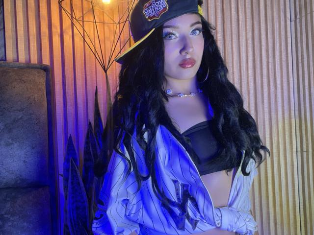 LillyTaylor with long black wavy hair, wearing a black cap, blue shirt open to show black bra, hoop earrings, and choker, blue and orange lighting, looking at camera.