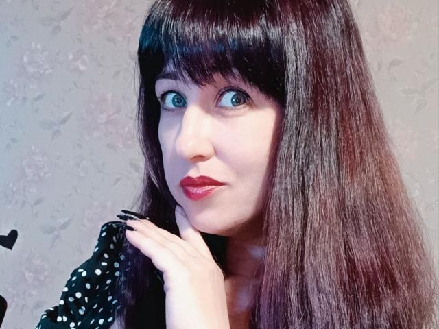JillSilvies has dark, shiny hair with bangs, blue eyes, and red lipstick. She's holding a black polka dot shirt, showing her white, smooth tits.