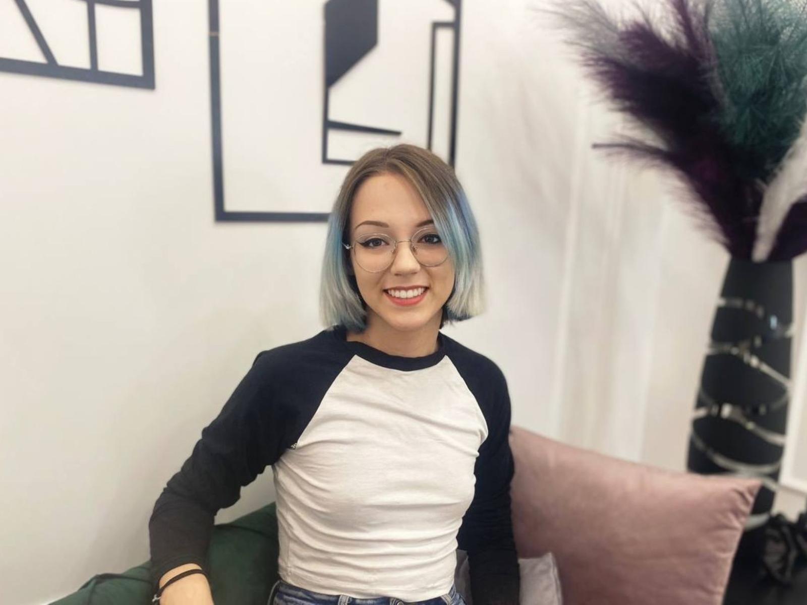 EnergyofLove, smiling with blue-tinted bob, wearing a black and white raglan shirt, sitting on a green couch, has small tits, cute, and attractive.