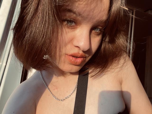 Ssimona with shoulder-length brown hair, blue eyes, and orange lipstick, wearing a black strap and silver chain, looking at the camera with slightly pouted lips.
