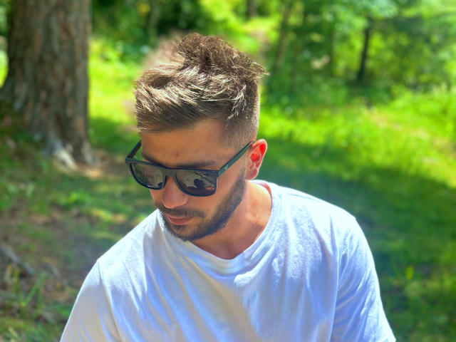 OnlyRoro with short brown hair, beard, black sunglasses, and white tee, looking down, standing in sunlit forest, greenery in background.