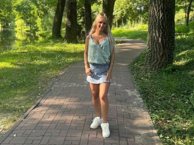 IsabelleEthan with blonde hair, wearing a loose green top, denim skirt, and white sneakers, standing on a brick path, trees in the background, looking down with a slight smile.