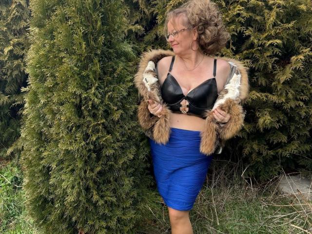 Vollbusige100dd-hot stands with curly brown hair, glasses, black bra, blue skirt, and fur jacket, holding jacket edges, showing her tits, against green bushes.