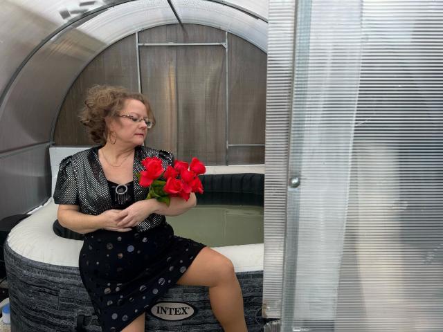 Vollbusige100dd-hot, middle-aged with curly brown hair, glasses, black polka-dot dress, holding red roses, sitting by a metallic hot tub, showing her ass.