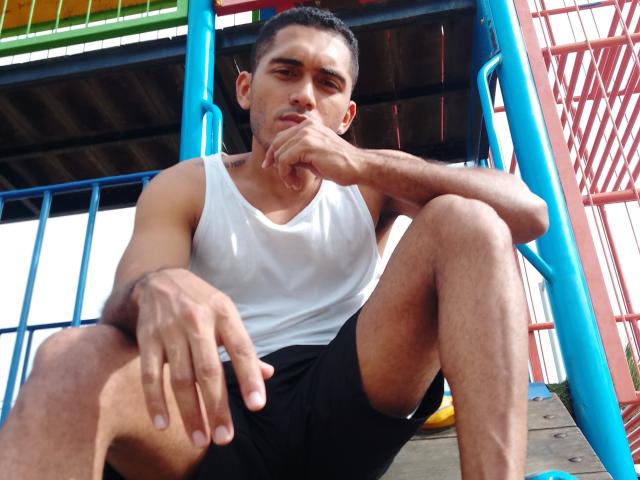 DereckStorm, with short dark hair, wearing a white tank top and black shorts, sits on a blue and red metal playground structure, resting his chin on his hand.