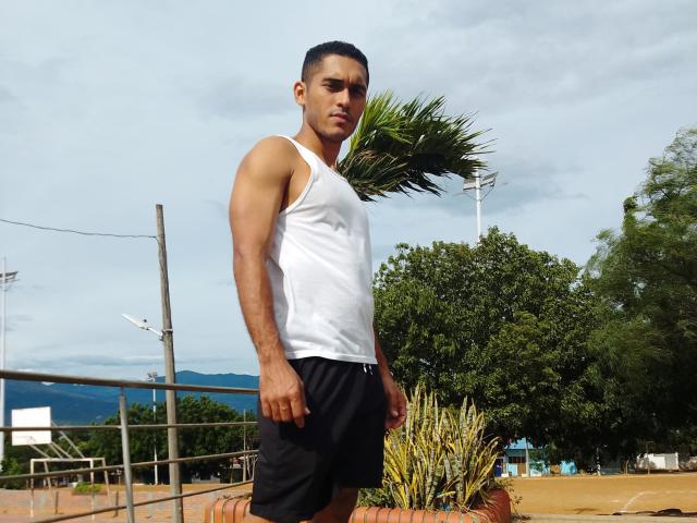 DereckStorm, muscular Latino guy with short black hair, white tank top, black shorts, standing outside, palm tree in background, serious expression, sunny day, green trees, light blue sky.