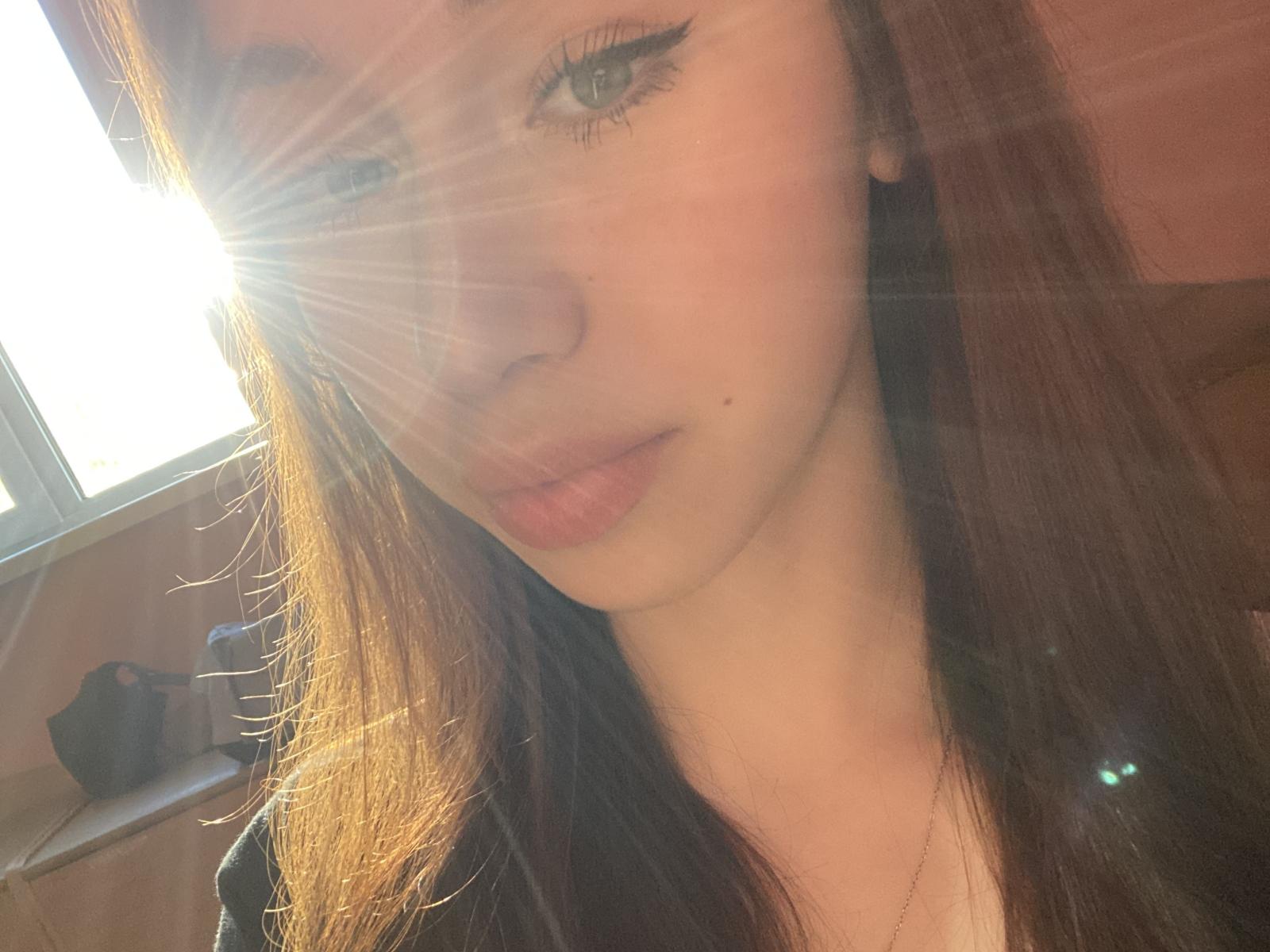 MuaShimur's face with sun flares, light brown hair, subtle makeup, and pink lips. Her eyeliner is sharp, and she has a small beauty mark on her left cheek.