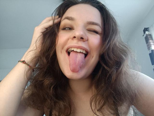 Dreia has wavy brown hair, a cute face, and is sticking out her tongue with one eye closed. She's wearing a bracelet and a tank top, looking playful.